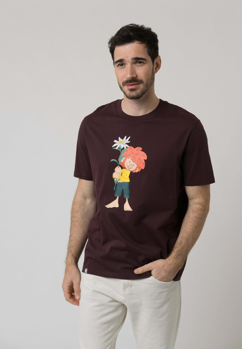 Man with short dark hair and beard wearing a dark t-shirt with a cartoon character holding a large white flower, white pants, hand in pocket.
