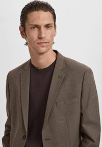 Bruine gestructureerde blazer over een donkere trui, met geknipte revers en een enkele borstzak, met een gladde afwerking en een getailleerde pasvorm.