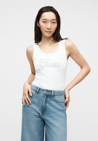Jeune femme aux cheveux lisses et foncés portant un débardeur blanc avec le texte "Guess" et un jean délavé bleu clair ample, posant les mains sur les hanches.