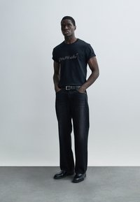 Μαύρο βαμβακερό t-shirt με κοντά μανίκια και τυπωμένο λογότυπο, σε συνδυασμό με σκούρο ντένιμ παντελόνι και μαύρα παπούτσια, με ουδέτερο φόντο.