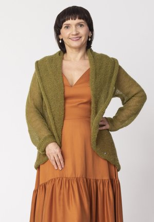 Kobieta o krótkich ciemnych włosach w pomarańczowej sukience i luźnym oliwkowozielonym dzierganym kardiganie, uśmiechająca się z jedną ręką na biodrze.