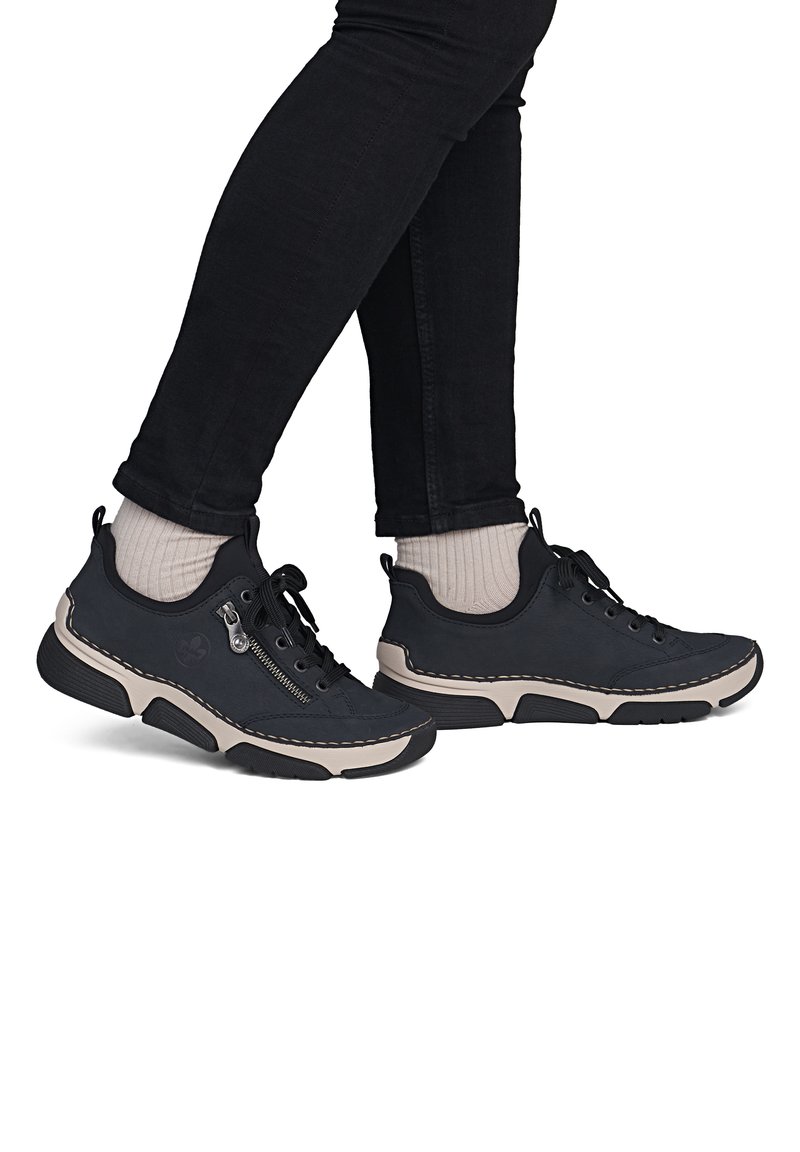Pieds en baskets noires avec semelles beiges, chaussettes beiges et pantalons noirs sur fond blanc, un pied légèrement levé.