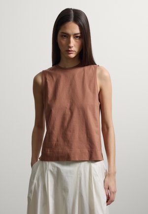 Vrouw met steil donker haar die een mouwloze bruine top en een witte geplooide rok draagt, staand tegen een effen lichte achtergrond.