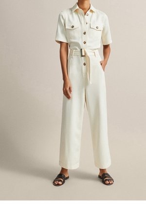 Personne portant une combinaison crème à manches courtes boutonnée avec ceinture à la taille et des sandales noires à bouts ouverts sur fond beige.
