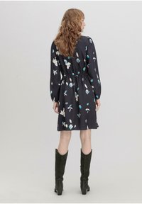 Schwarzes Kleid mit floralen Mustern in Pastellfarben, tailliert, mit langen Ärmeln und einem ausgestellten Rock, kombiniert mit grünen knielangen Stiefeln.