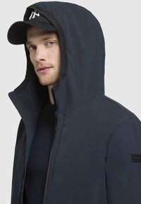 Marineblå hættejakke lavet af glat stof, med lynlås og et brandmærke på ærmet. Sort baseballcap under hætten.