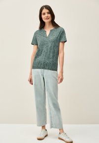 Grøn kortærmet top med et geometrisk mønster og v-hals, parret med lyseblå brede jeans og hvide sneakers. Enkel design.