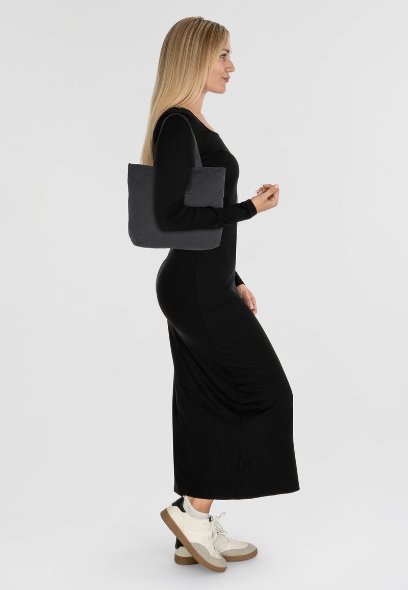 Bolso tote negro acolchado con dos asas. Una modelo lleva un vestido largo negro y zapatillas blancas, mostrando la textura suave y el diseño del bolso.