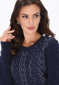 Tummansininen neulottu villapaita, jossa on koristeellisia strasseja lehden muotoisessa kuviossa ja helmiaksentteja olkapäillä. Pehmeä tekstuuri, pyöreä kaula-aukko.