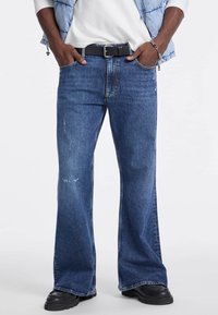 Jeans in denim blu a zampa d'elefante con una finitura sbiadita, dotati di tasche frontali, chiusura con bottone e una cintura nera indossata alta in vita.
