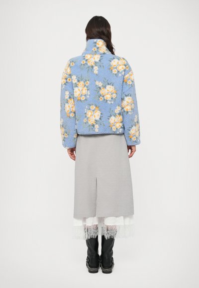 Kohev sinine fliisjope kollaste ja valgete lillemustriga, kõrge krae ja lõdva lõikega, sobitatud halli seeliku ja mustade saapadega.