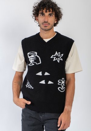 Homme aux cheveux bouclés portant un gilet en tricot noir avec des motifs abstraits blancs sur une chemise beige à manches courtes et un jean foncé.