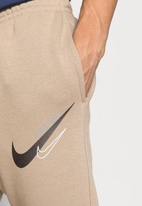 Béžové tepláky s hladkým povrchem, s černobílým logem Nike a textem "Just do it" v stříbře na boční kapse.