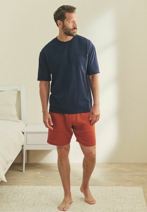 Navy short-sleeve t-shirt with a pocket paired with textured orange shorts, set against a light-colored room with wooden flooring.