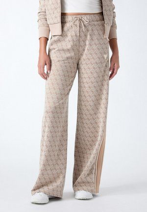 Femme portant un pantalon large beige à motif avec bande latérale et baskets blanches, mains détendues le long du corps.