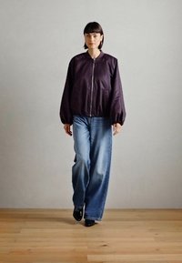 Giacca bomber viola scuro con chiusura a zip, abbinata a jeans a gamba larga blu chiaro e scarpe nere. Tessuto liscio, design informale.