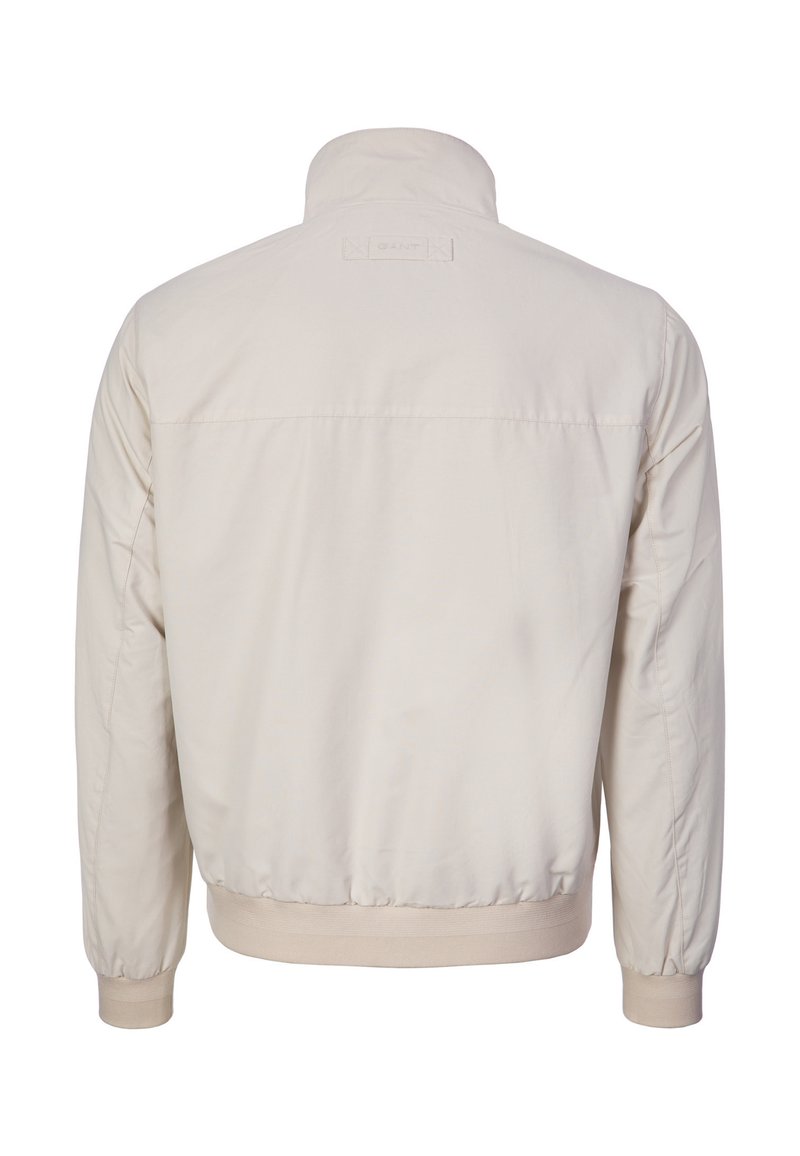 Giacca casual beige chiaro con polsini e orlo a costine, collo alla coreana e etichetta del marchio cucita sulla parte superiore della schiena, vista da dietro.