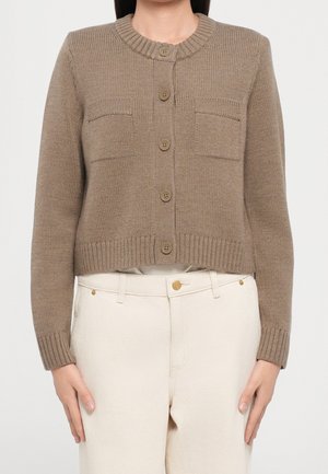 Femme portant un cardigan en maille marron boutonné avec deux poches poitrine et un pantalon crème taille haute sur fond uni.