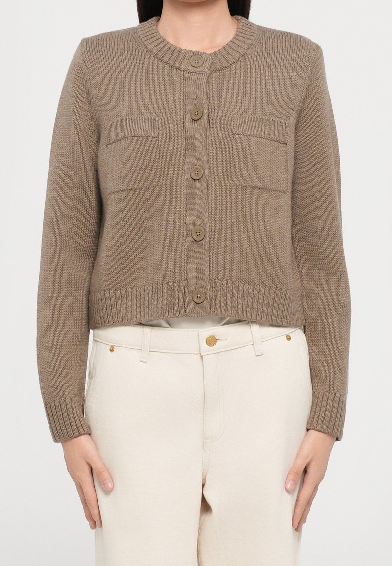 Femme portant un cardigan en maille marron boutonné avec deux poches poitrine et un pantalon crème taille haute sur fond uni.
