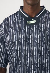 Mann trägt ein marineblaues Hemd mit weißem geometrischen Muster und schwarzem Kragen, das ein hellgrünes Puma-Logo und -Text aufweist.