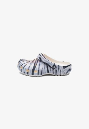 Zueco estilo slip-on con patrón tie-dye en blanco y negro, con orificios de ventilación y correa ajustable en el talón, detalle del logo lateral visible.
