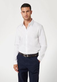 Camisa blanca de manga larga con cuello abotonado y un pequeño logo en el pecho, combinada con pantalones azul marino oscuros y un cinturón marrón con hebilla de metal.