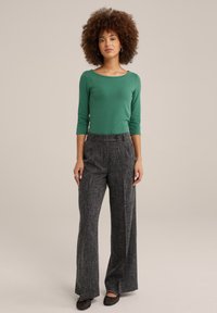 Grön långärmad topp med rund halsringning, kombinerad med högmidjade byxor i grått tweed med vida ben och svarta platta skor.