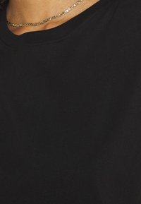 T-shirt en coton noir avec col rond et manches courtes, présentant des détails de couture soignés et une texture lisse. Collier chaîne discret visible.