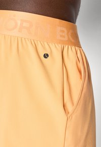 Light orange athletic shorts with an elastic waistband featuring embossed text. Includes a side pocket and a small black logo.