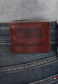 Brun läderlapp med präglad "TOMMY JEANS" och "AN AMERICAN ORIGINAL" på mörk denim. Synlig söm längs midjebandet.