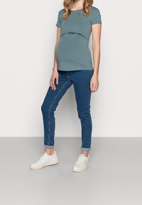 Těhotenské tričko v tlumené modrozelené barvě s vrstevnatým designem, spárované s úzkými modrými džíny a bílými teniskami. Jednoduchý a moderní styl.
