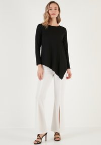 Top nero a maniche lunghe con un design a coste testurizzate e un orlo asimmetrico, abbinato a pantaloni bianchi a palazzo con spacchi laterali. Tacchi neri con cinturini.