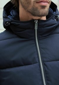 Jachetă puffer de culoare albastru închis, cu guler înalt și glugă, având un închidere cu fermoar argintie și un material texturat. Fără logo-uri sau modele vizibile.