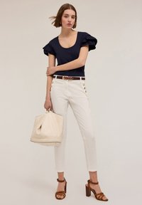 Haut froncé bleu marine, pantalon blanc ajusté avec des boutons dorés, ceinture marron, sac beige et sandales à talons marron avec des lanières.