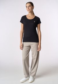 T-shirt nera a maniche corte, scollo rotondo, tessuto morbido, accentuazione del logo dorato sul petto, abbinata a pantaloni beige chiaro e sneakers bianche.