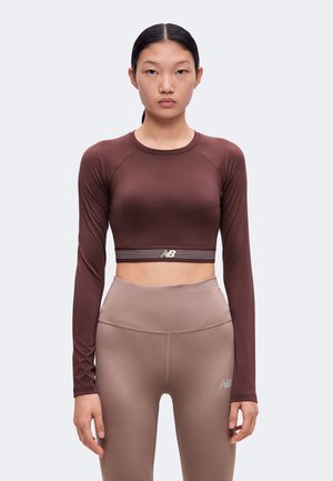Femme portant un crop top marron ajusté à manches longues et des leggings beige taille haute, debout devant un fond clair uni.