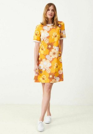 Kvinde iført en kortærmet, knælængde kjole med stort gult, orange og hvidt blomsterprint, parret med hvide sneakers, stående mod en hvid baggrund.