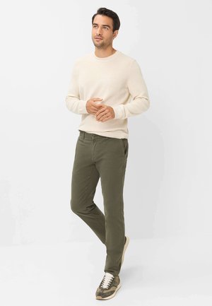 Mann trägt einen cremefarbenen Pullover, olivgrüne Hose und Camouflage-Sneakers, steht vor einem einfarbigen hellen Hintergrund und schaut leicht nach oben.