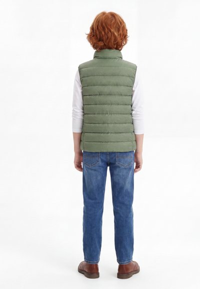 Παιδί με κόκκινα σγουρά μαλλιά φοράει πράσινο αφράτο γιλέκο, λευκό μακρυμάνικο πουκάμισο, μπλε τζιν και καφέ παπούτσια, στέκεται με την πλάτη σε λευκό φόντο.