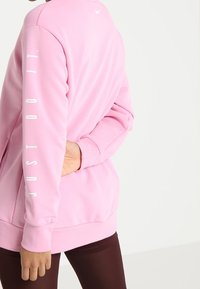 Sudadera rosa de ajuste holgado y mangas largas. Presenta el texto "just do it" en blanco en la manga izquierda y un pequeño logo en la parte trasera.