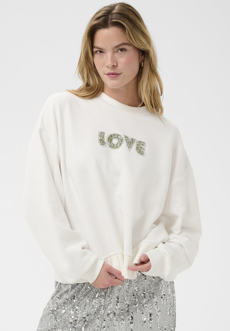 Femme aux cheveux blonds portant un sweat-shirt blanc avec "LOVE" en strass et une jupe à paillettes argentées, posant devant un fond uni.