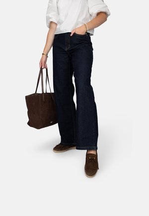 Donkerblauwe high-waisted jeans met een wijde pijp, gecombineerd met een witte blouse en een bruin suède tas. Bruine mocassinschoenen met franjes.