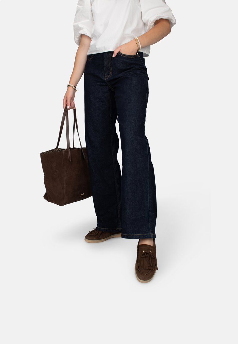 Jeans de tiro alto de color azul oscuro con pierna ancha, combinados con una blusa blanca y un bolso de ante marrón. Zapatos estilo mocasín marrones con flecos.