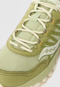 Világoszöld sneaker, hálós és velúr textúrákkal, bézs fűzőkkel, fényvisszaverő részletekkel és egy egyedi logóval az oldalon.