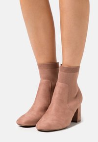 Stivaletti in suede rosa polveroso alti fino alla caviglia, con risvolti a costa in lavorazione a maglia e tacchi a blocco, indossati su gambe scoperte.