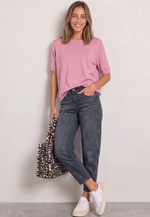 Femme souriante, portant un t-shirt rose à manches courtes, un jean gris foncé, des baskets blanches, tenant un sac fourre-tout imprimé léopard, debout contre un mur clair.