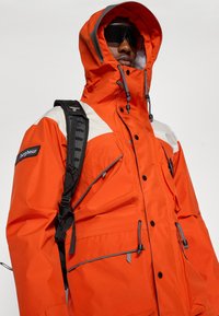 Bright orange waterproof jacket with gray accents, adjustable hood, multiple pockets, and a secure zipping system. Features a backpack strap.