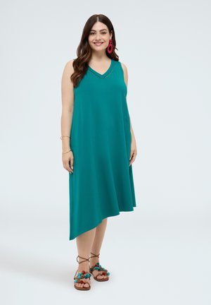 Lachende vrouw die een mouwloze asymmetrische teal jurk draagt, grote rode oorbellen en bruine sandalen versierd met kleurrijke pompons, staand tegen een effen achtergrond.