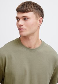 Tricou de culoare verde măslinie dintr-un material moale, cu un gât rotund, mâneci scurte și croială lejeră. Prezentă detalii discrete de cusătură.