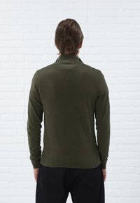 Pull en tricot vert olive avec un col montant et des manches longues, présentant des bords à côtes au niveau des poignets et de l'ourlet. Texture lisse sur un fond de briques blanches.